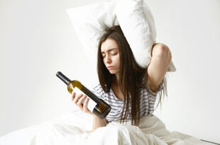 stressed unhappy woman with messy loose hair suffering from headache after party holding pillow empty bottle